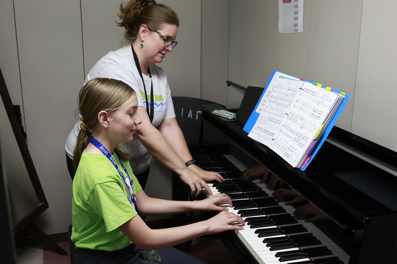 A student practicing piano in a practice room with a teacher.